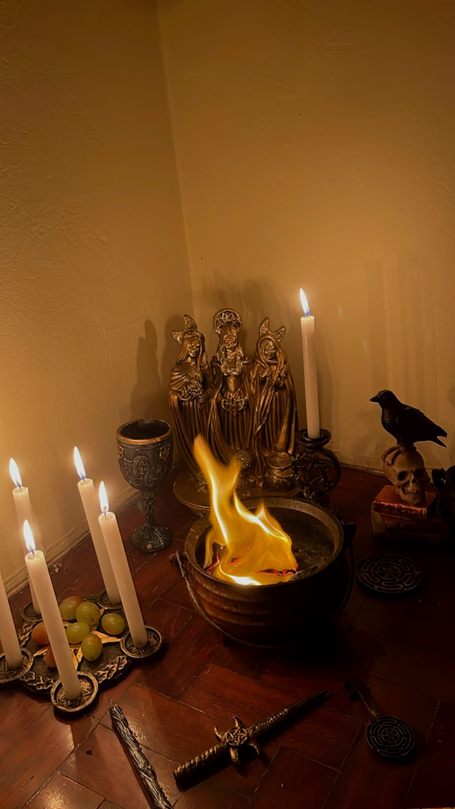 Caldeirão com fogo aceso, velas brancas, cálice, estátua dourada de três figuras e um corvo sobre uma caveira