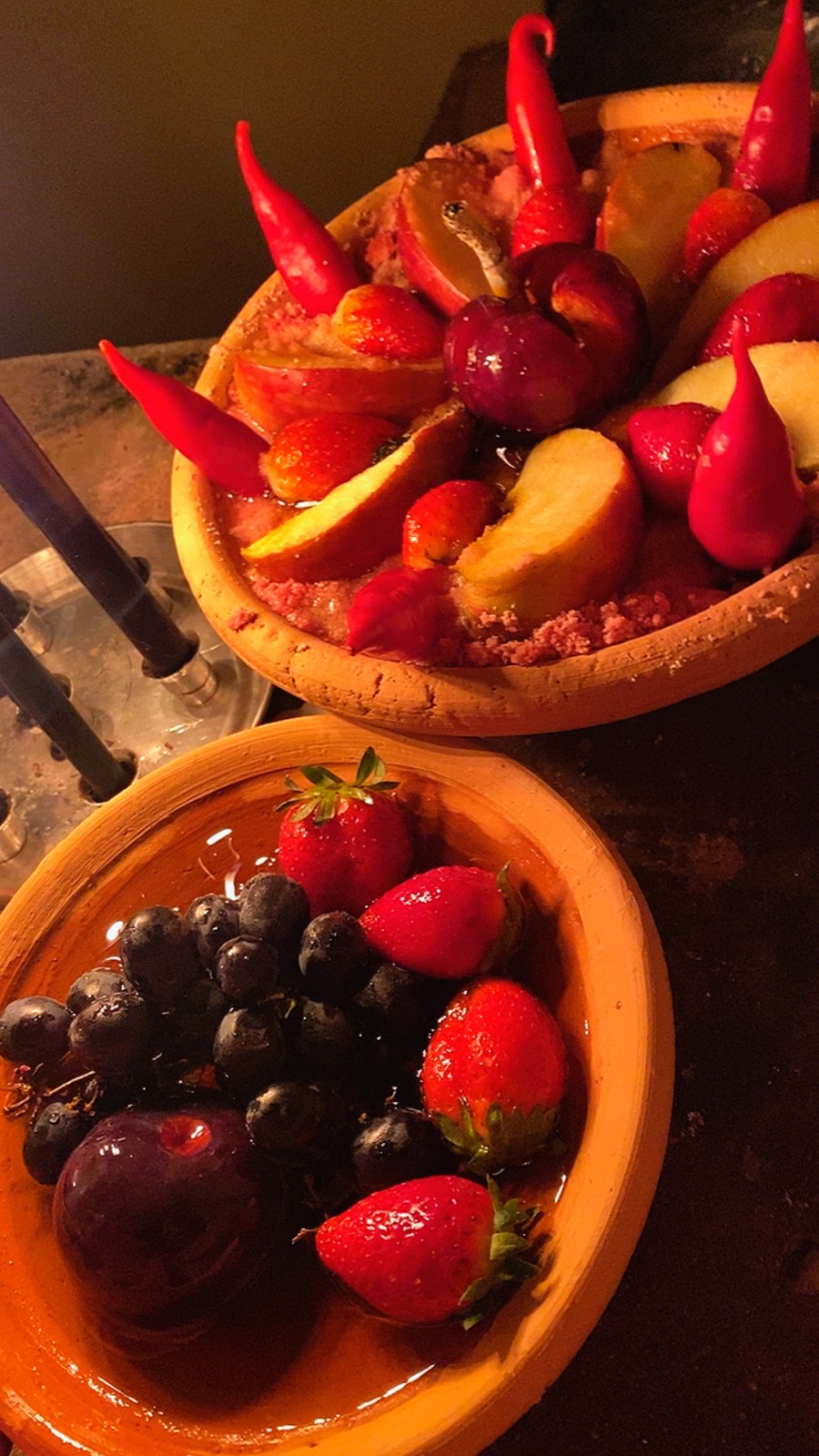 Dois pratos de barro com morangos, uvas, maçã fatiada, ameixa e pimentas vermelhas, com velas pretas ao lado