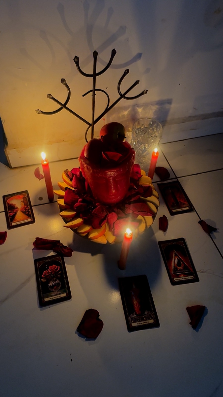 Vela vermelha grande cercada por pétalas de rosa e fatias de fruta, com cartas de tarot dispostas ao redor e três velas acesas